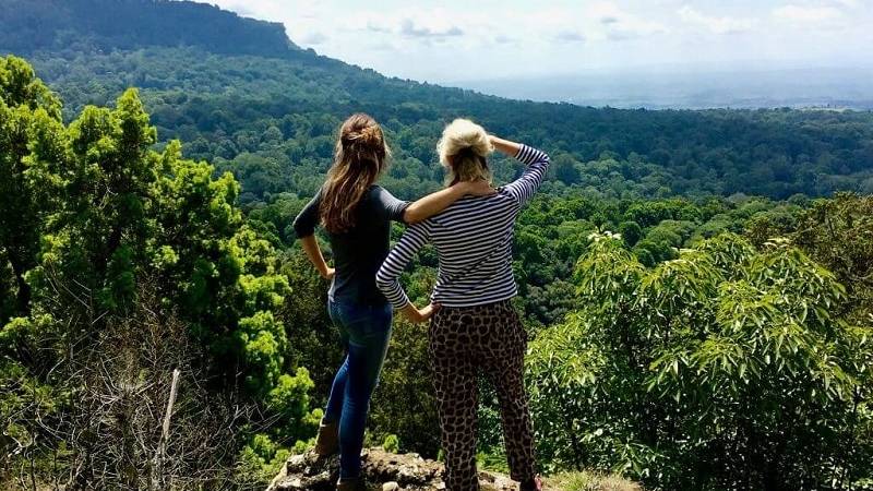 Two women stand on a rocky ledge overlooking a lush green forest and mountains, embracing as they take in the breathtaking Kenyan landscape—a perfect moment while travelling through Africa.