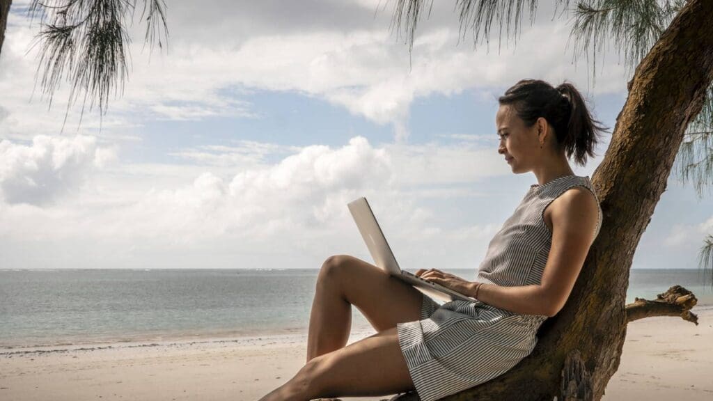 A woman sits on a tree trunk by the beach in Kenya, using a laptop. Wearing a sleeveless dress, she appears focused on her work. The ocean and sky are visible in the background, framed by tree branches—a perfect scene for travelling in Africa.