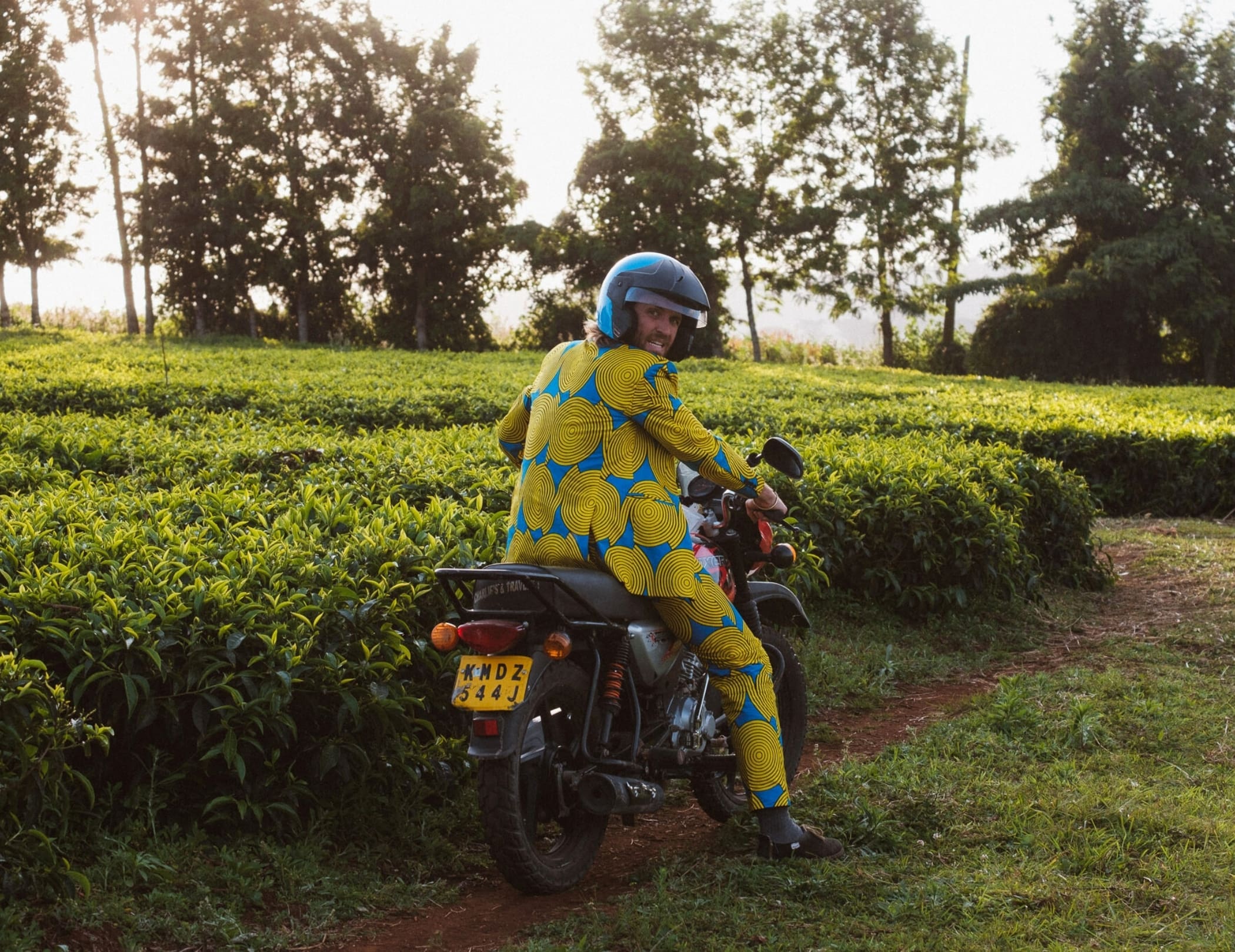 Een persoon in een felgeel en blauw pak met helm zit op een motorfiets op een onverhard pad naast weelderige groene velden, met bomen op de achtergrond - een inspirerende scène voor reizigers die Kenia verkennen.