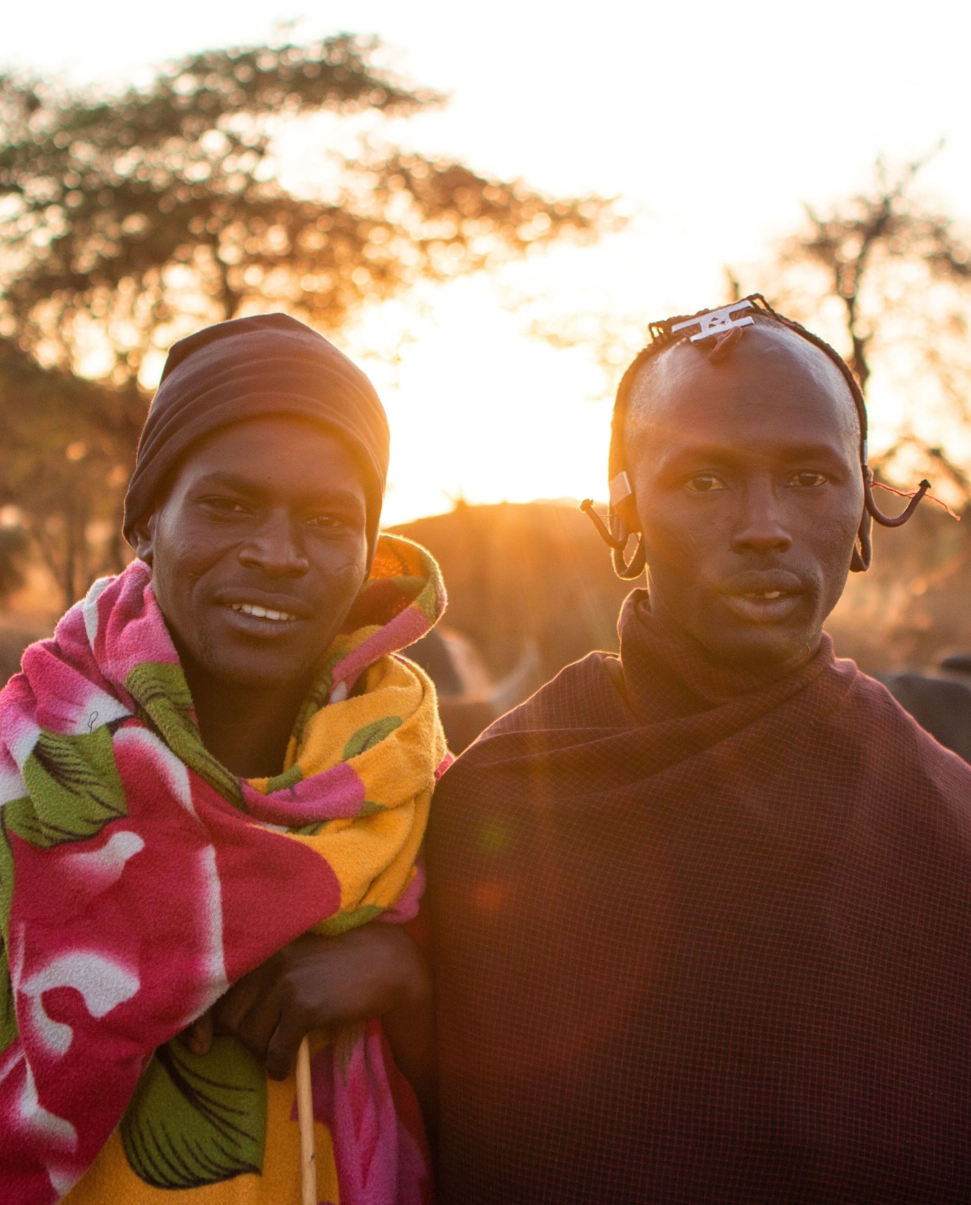 Twee mannen in kleurrijke traditionele kleding staan zij aan zij buiten bij zonsondergang, met bomen zacht vervaagd op de achtergrond. De ene man draagt een roze deken met patroon, de andere een donkere sjaal en oorbellen.