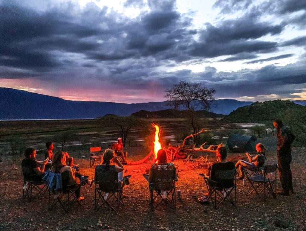 Een groep mensen zit in een cirkel van stoelen rond een kampvuur in de schemering, met dramatische wolken en een kleurrijke zonsondergang op de achtergrond, omringd door open wildernis en heuvels.