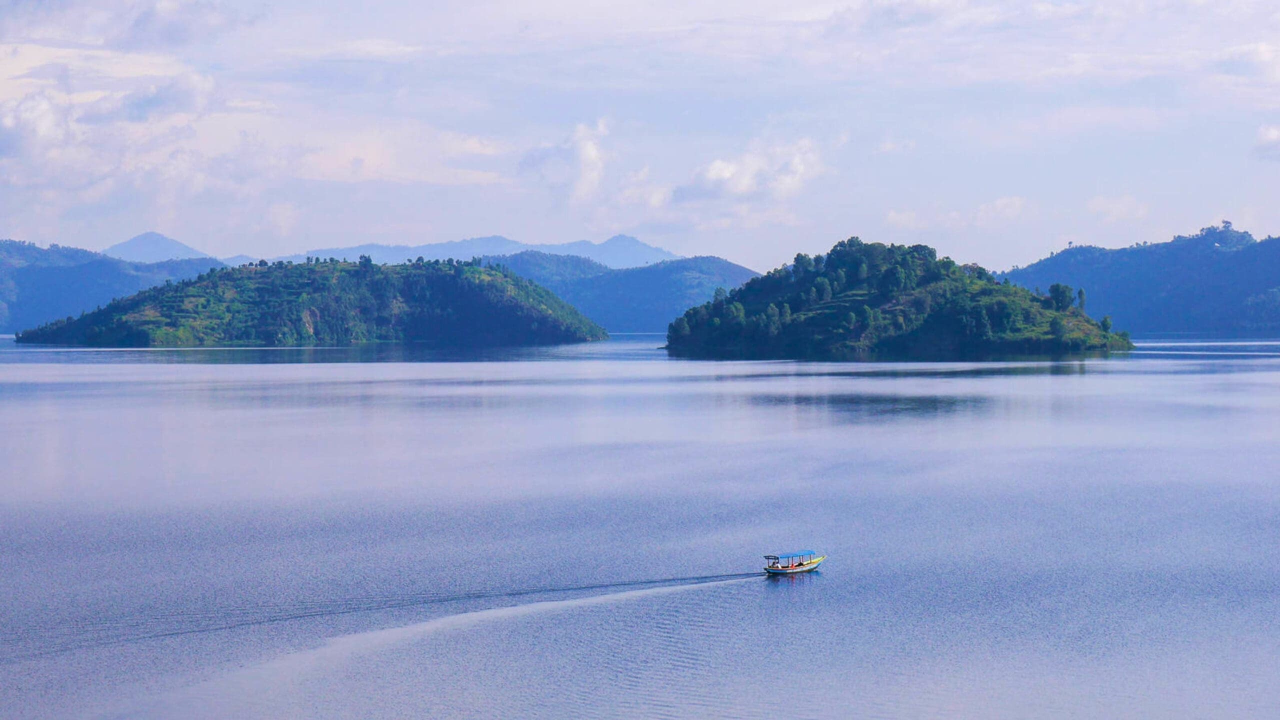 Een klein bootje vaart over een kalm blauw meer met twee groene, heuvelachtige eilanden op de achtergrond en bergen in de verte onder een gedeeltelijk bewolkte hemel.