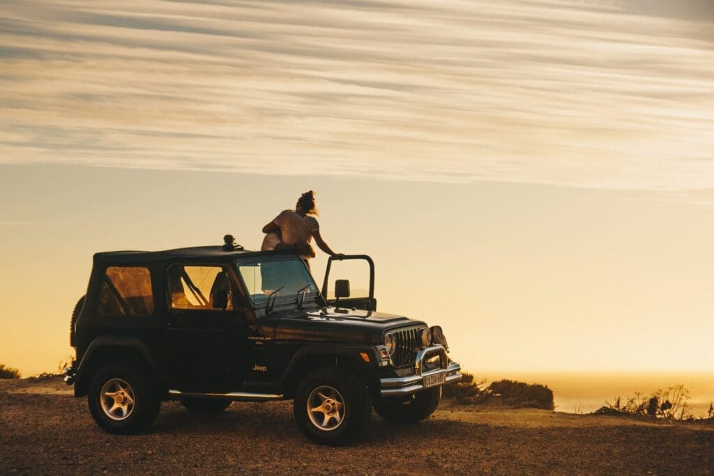 A person sits on the roof of a black Jeep parked on a dirt road in South Africa, gazing at a golden sunset with wispy clouds. The open landscape evokes a sense of adventure and freedom, perfect for anyone who loves travelling.