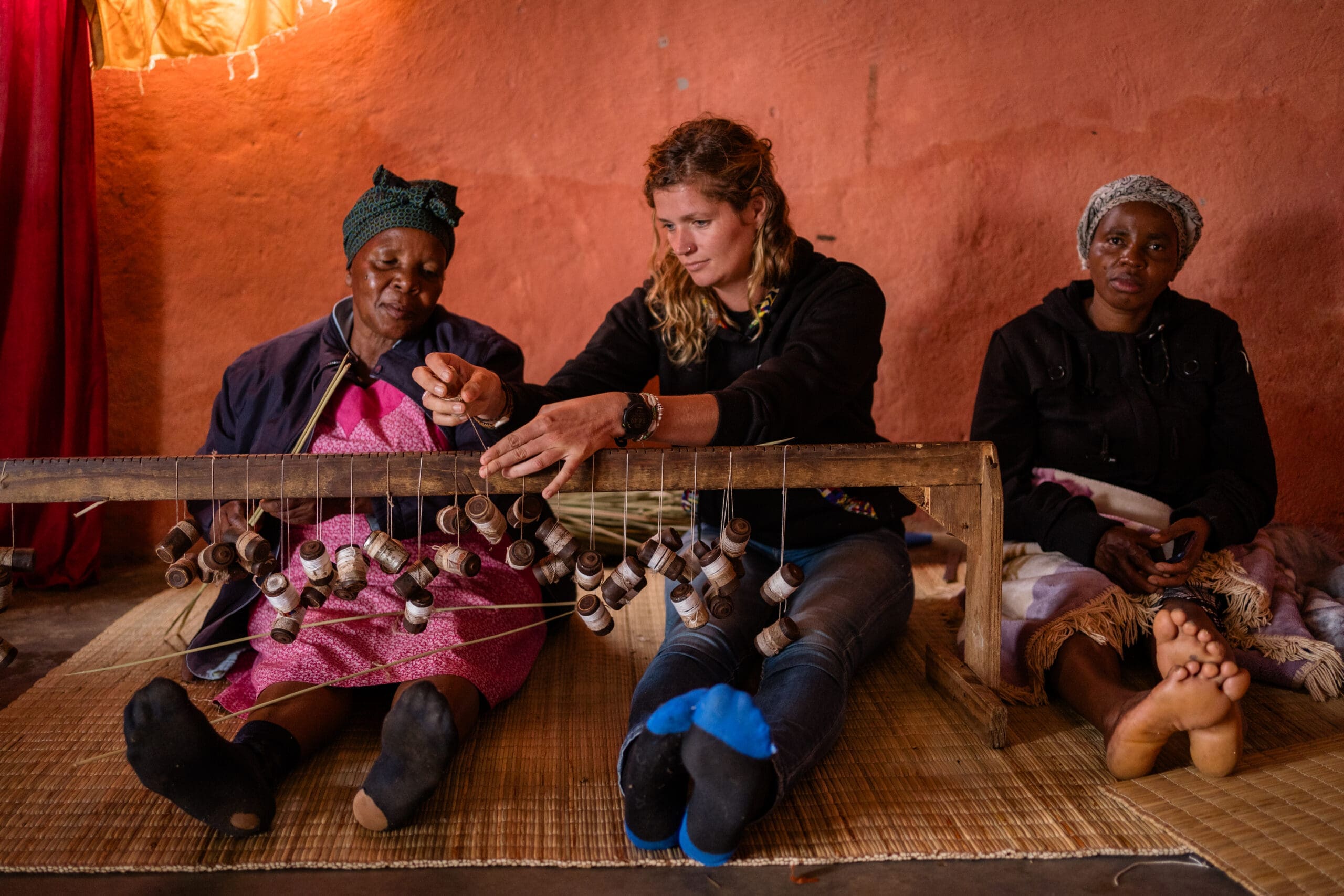 Three women in eSwatini sit on mats indoors, two weaving intricate patterns with a wooden loom while the third observes. The reddish-brown wall behind them adds warmth to this authentic glimpse of traditional life in Africa.