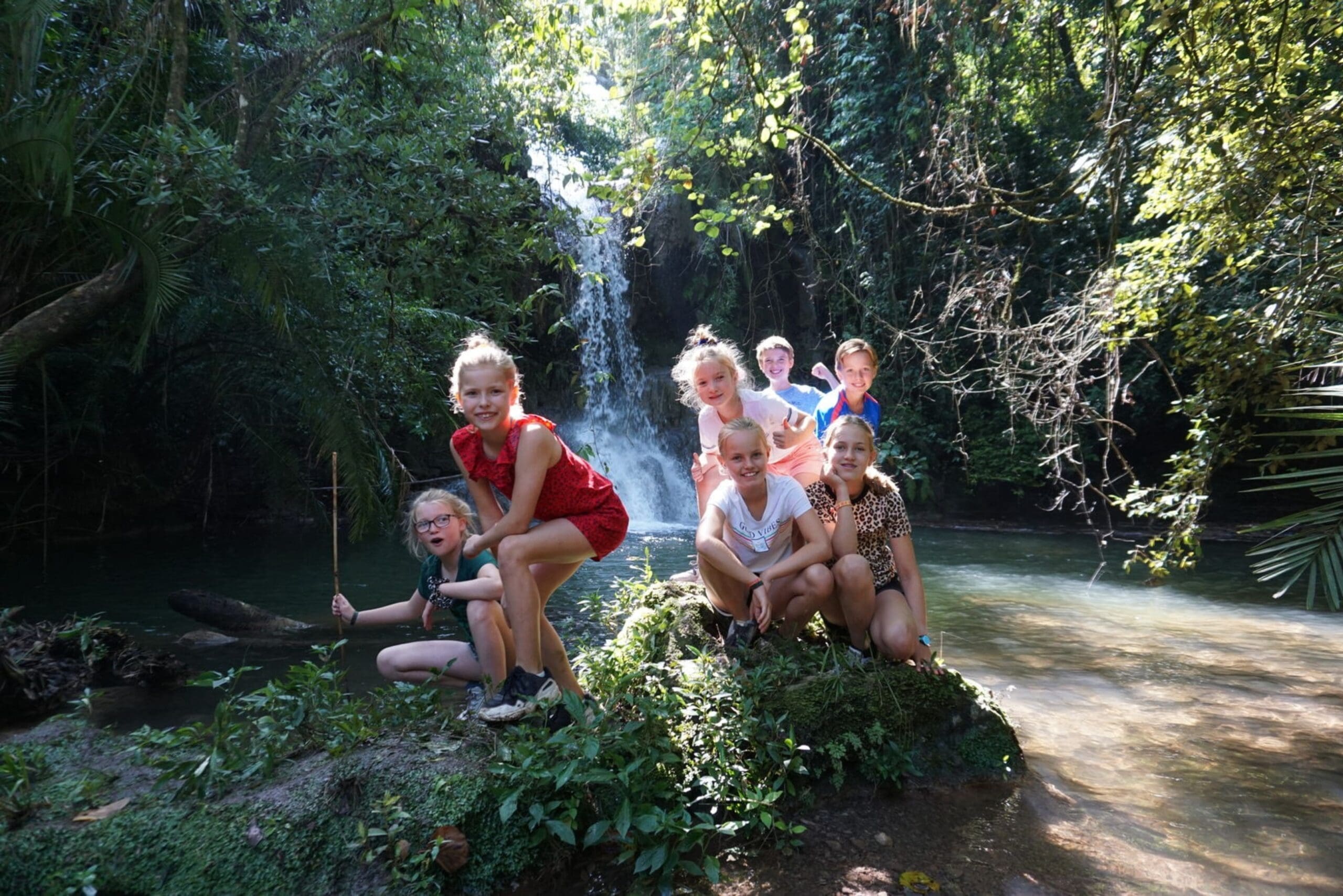 A group of children pose together on a mossy rock in front of a lush, green waterfall surrounded by trees and water, smiling and enjoying the sunny outdoor setting.