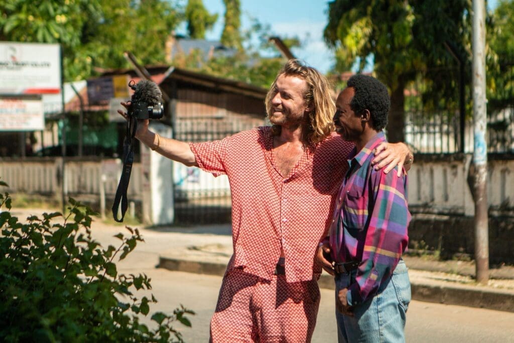 Two men stand outdoors on a sunny day in Tanzania. One, wearing a red patterned outfit, holds a camera with a microphone and puts his arm around the other man in a purple plaid shirt. They both smile and look at the camera, capturing their travelling adventure.