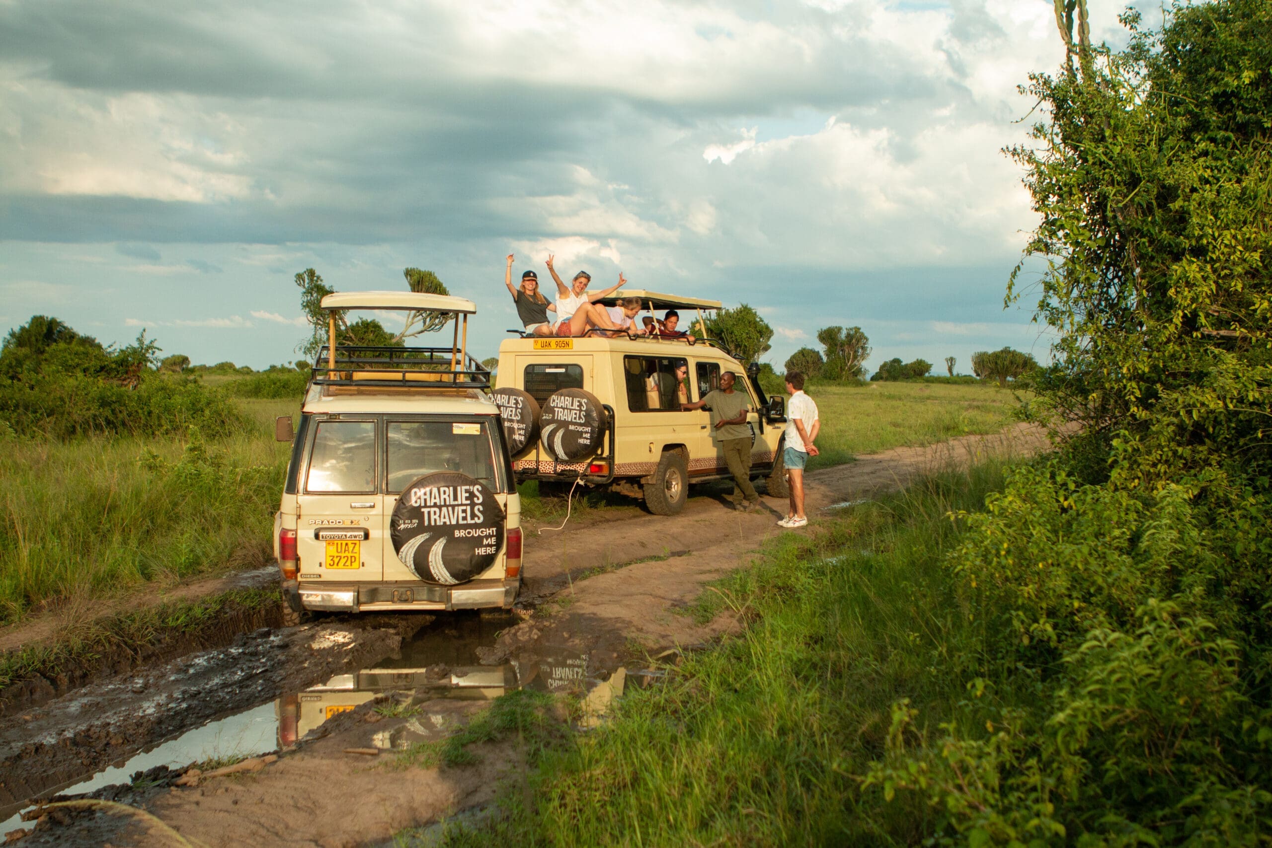 Two safari vehicles on a muddy dirt road in Uganda’s lush green landscape. Several people wave from the open roof of one vehicle, while another stands outside taking a photo. The sky is partly cloudy—an unforgettable Africa travelling experience.