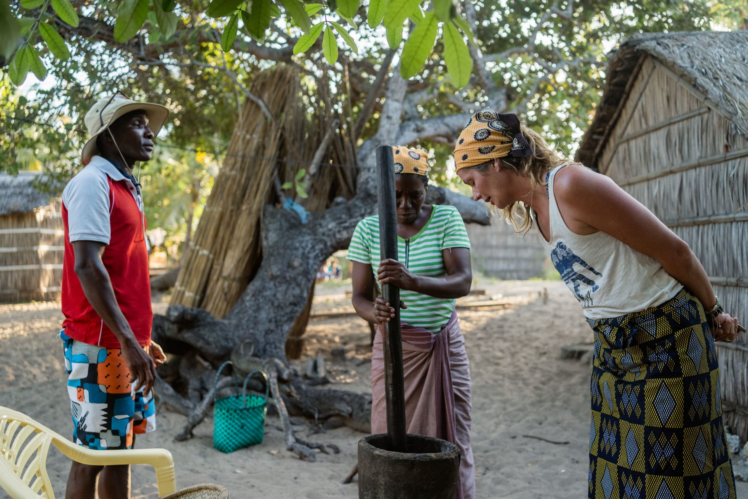 Drie mensen staan buiten in Mozambique bij traditionele hutten; een vrouw in het midden stampt voedsel in een vijzel, terwijl een andere vrouw aandachtig toekijkt en een man naast hen staat, allemaal onder lommerrijke Afrika-bomen.
