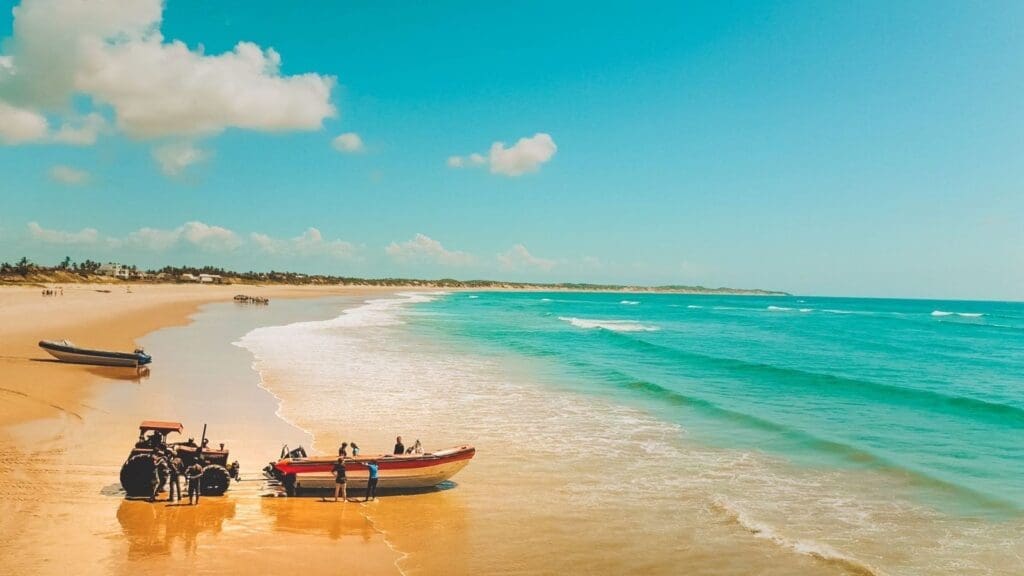 Een groep mensen staat naast kleurrijke boten op een zandstrand in Mozambique met helder turquoise water en zachte golven onder een heldere, zonnige Afrikaanse hemel. In de verte zijn bomen en meer mensen te zien langs de kustlijn.