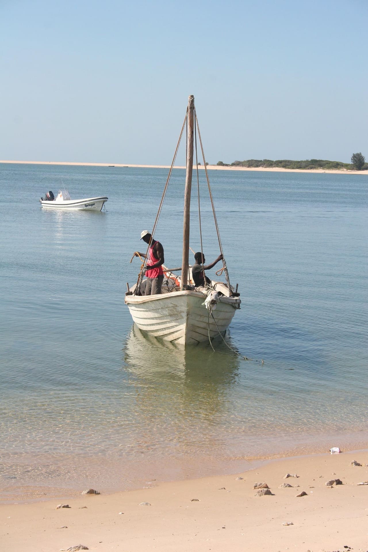 Two people sit in a small wooden sailboat near the sandy shore of calm, clear water in Mozambique. Another boat floats in the distance and lush African vegetation lines the horizon under a clear sky—perfect for travelling and exploring nature.