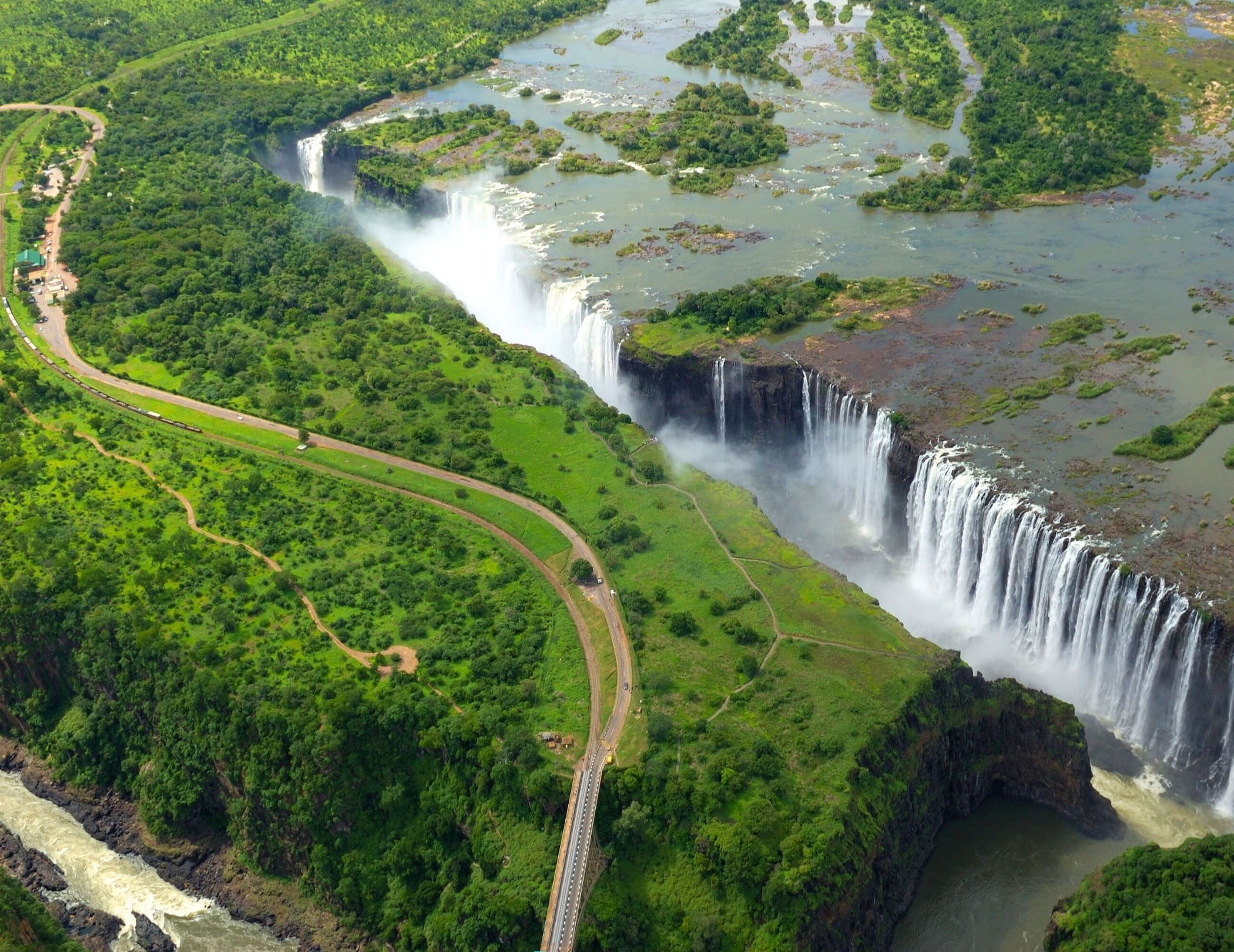 Luchtfoto van de Victoria Watervallen aan de rand van Botswana, met water dat over kliffen in een kloof stort, omringd door weelderige groene vegetatie en een kronkelige weg met een brug over de rivier in Afrika.