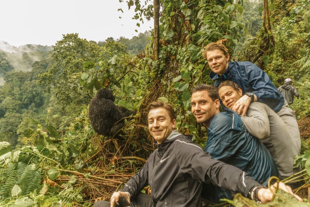 Vier reizigers in outdooruitrusting hurken en glimlachen voor een foto in een weelderig, groen bos in Afrika. Een gorilla zit vlakbij met zijn rug naar de camera, terwijl mistige bomen de achtergrond vullen, en de geest van reizen in het wild vastleggen.