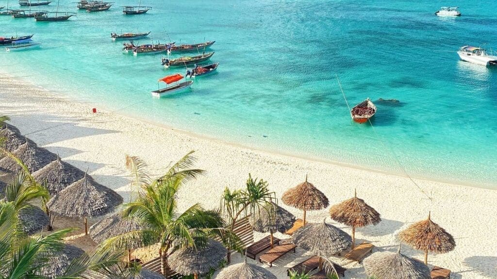 Thatched umbrellas and lounge chairs line a white sandy beach beside turquoise water. Several small boats float near the shore, with palm trees adding a tropical feel to the vibrant seaside scene.