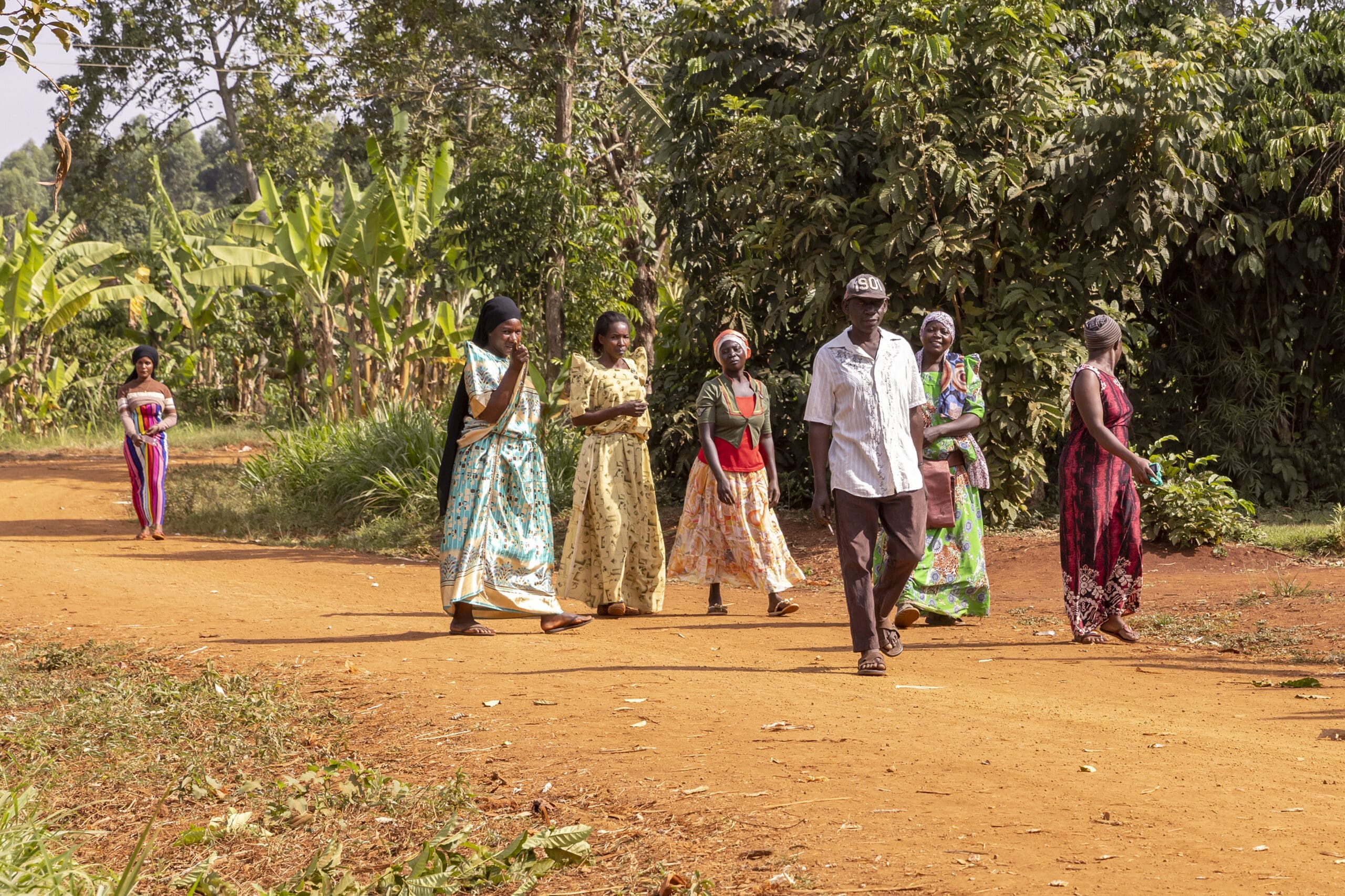 A group of people, mostly women in colorful dresses and one man in a white shirt, walk along a dirt path surrounded by green plants and trees in a rural setting. Another person stands further back on the path.