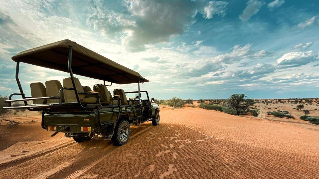 Een open safarivoertuig met lege stoelen staat geparkeerd op het roodachtige zand van Botswana onder een gedeeltelijk bewolkte hemel, met uitzicht op een uitgestrekt, droog Afrika-landschap dat bezaaid is met verspreide struiken en bomen.