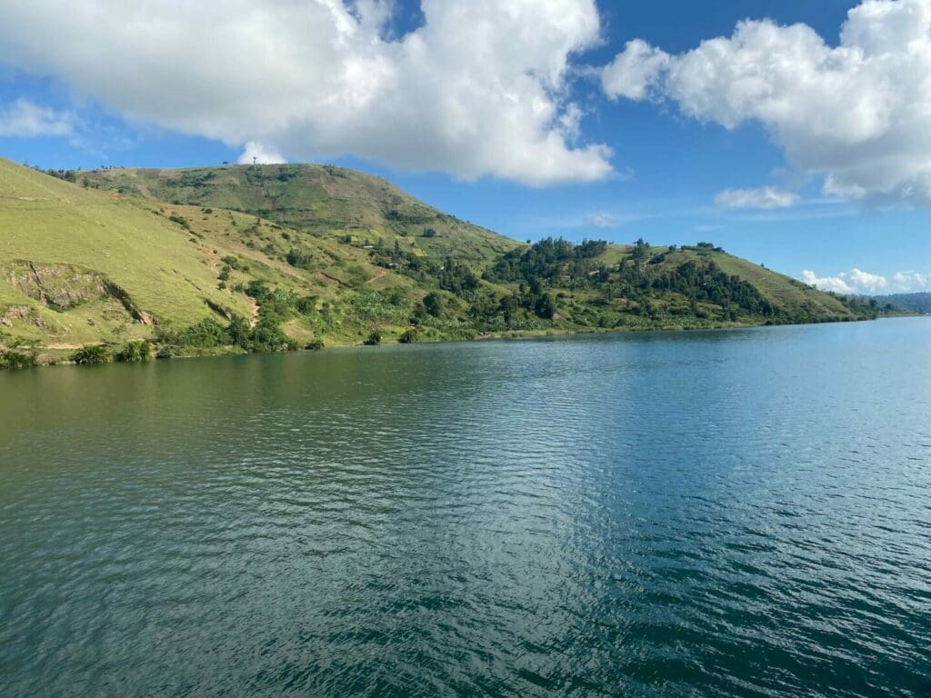 Een kalm blauw meer in Rwanda strekt zich uit onder een halfbewolkte hemel, met groene heuvels aan de overkant en weerspiegelingen die glinsteren op het wateroppervlak - een rustig tafereel in het hart van Afrika.