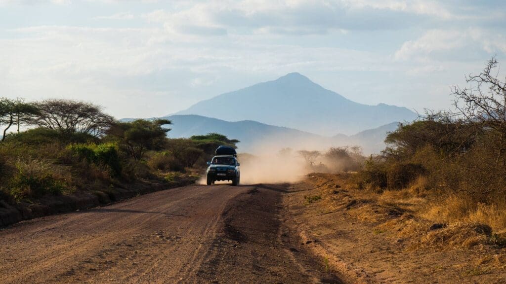 A vehicle drives on a dusty dirt road through a dry, rugged landscape in Tanzania, with scattered trees and a distant mountain rising under a partly cloudy sky—a true travelling adventure in Africa.