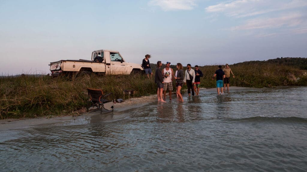 A group of people stand and talk by the water's edge near a grassy bank in South Africa, with a white pickup truck parked nearby. A folding chair and cooler sit on the sand as dusk sets in, perfect for travellers seeking relaxation.