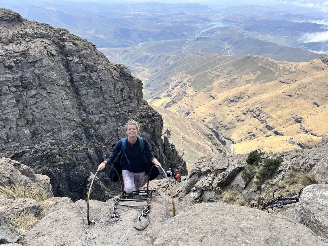 Een persoon met licht haar beklimt een metalen ladder die is bevestigd aan een rotsachtige berg in Zuid Afrika, omgeven door steile kliffen en weidse uitzichten over de vallei onder een bewolkte hemel-een adembenemende reiservaring.