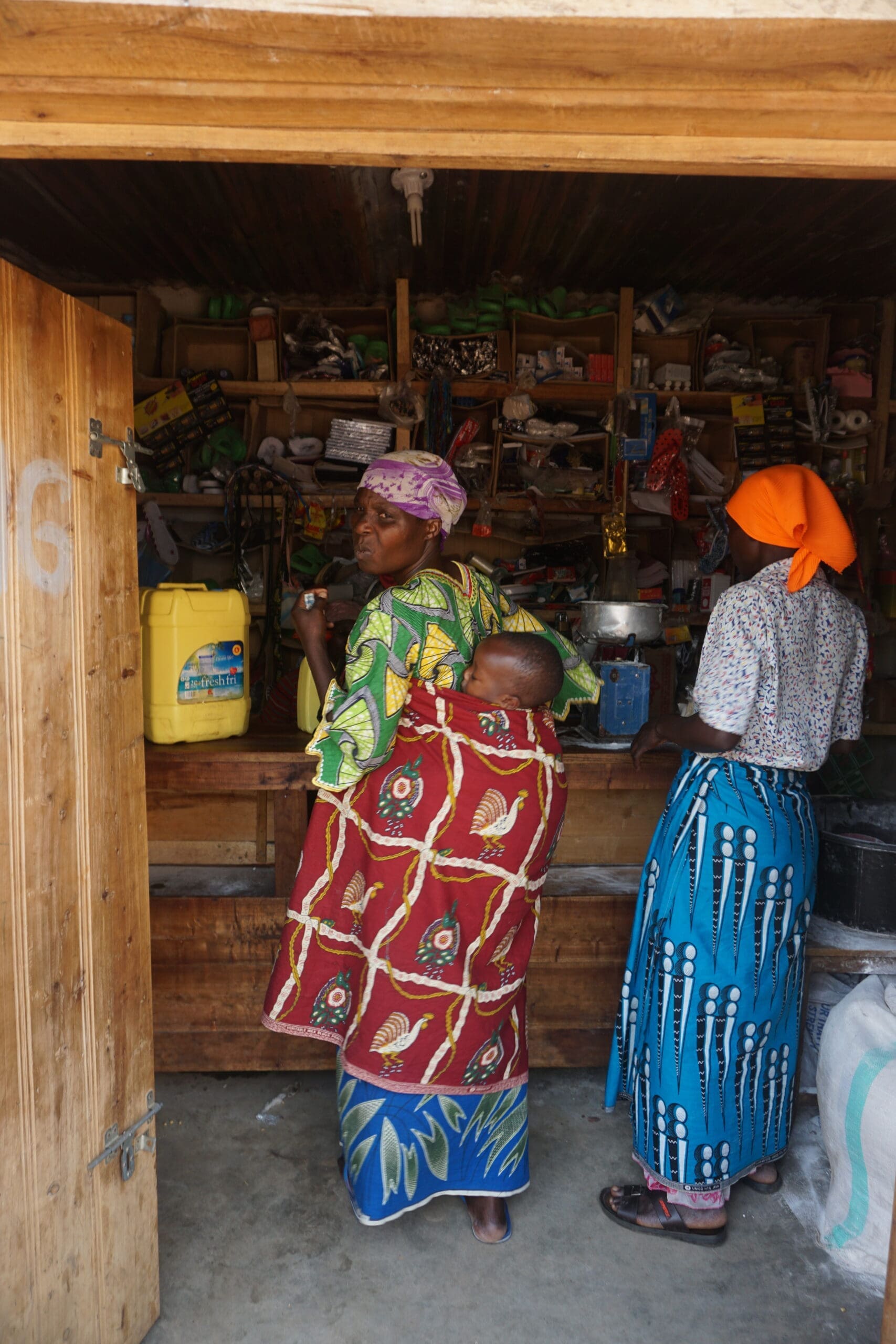 Two women in colorful traditional clothing stand in a small, wooden shop filled with various items. One woman carries a baby on her back, wrapped in a patterned cloth, while the other faces the shelves.