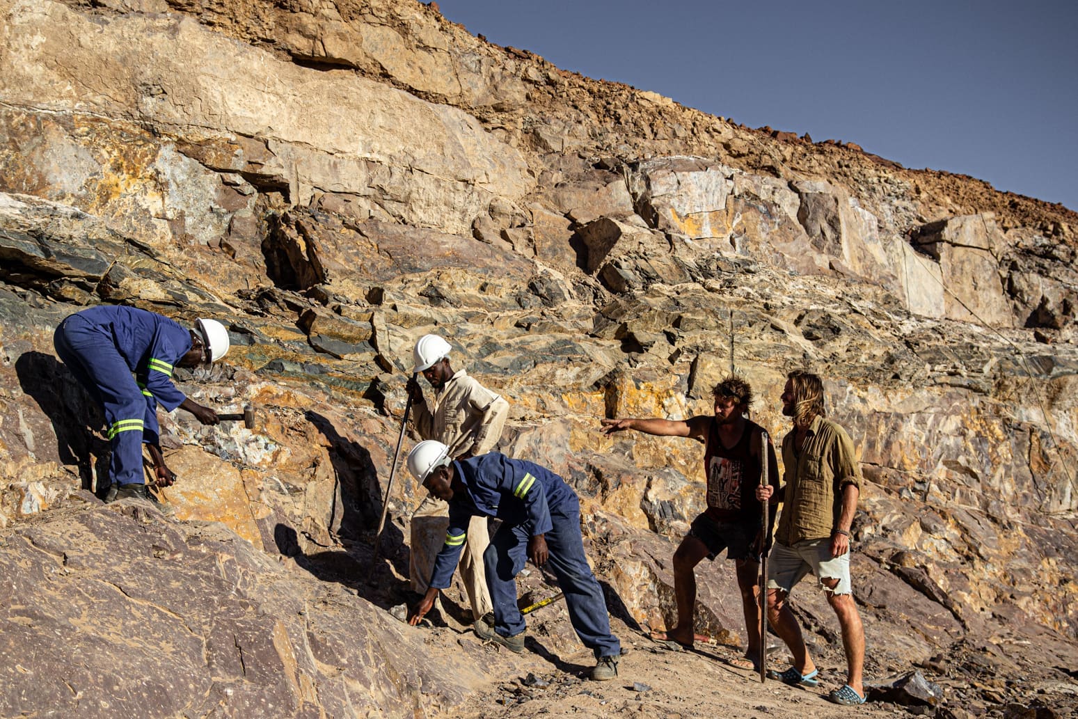 Four people wearing safety helmets and uniforms examine rocks on a rocky slope, while two others in casual clothing observe and point at the site. The scene appears to be an outdoor geological or archaeological expedition.