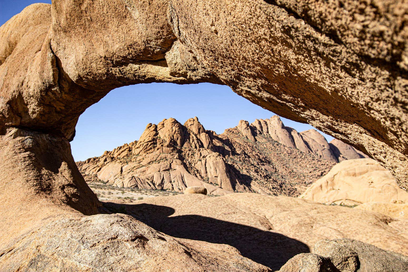 Een stenen boog omlijst ruige, zonovergoten bergen en rotsachtig terrein onder een strakblauwe hemel in een woestijnlandschap.