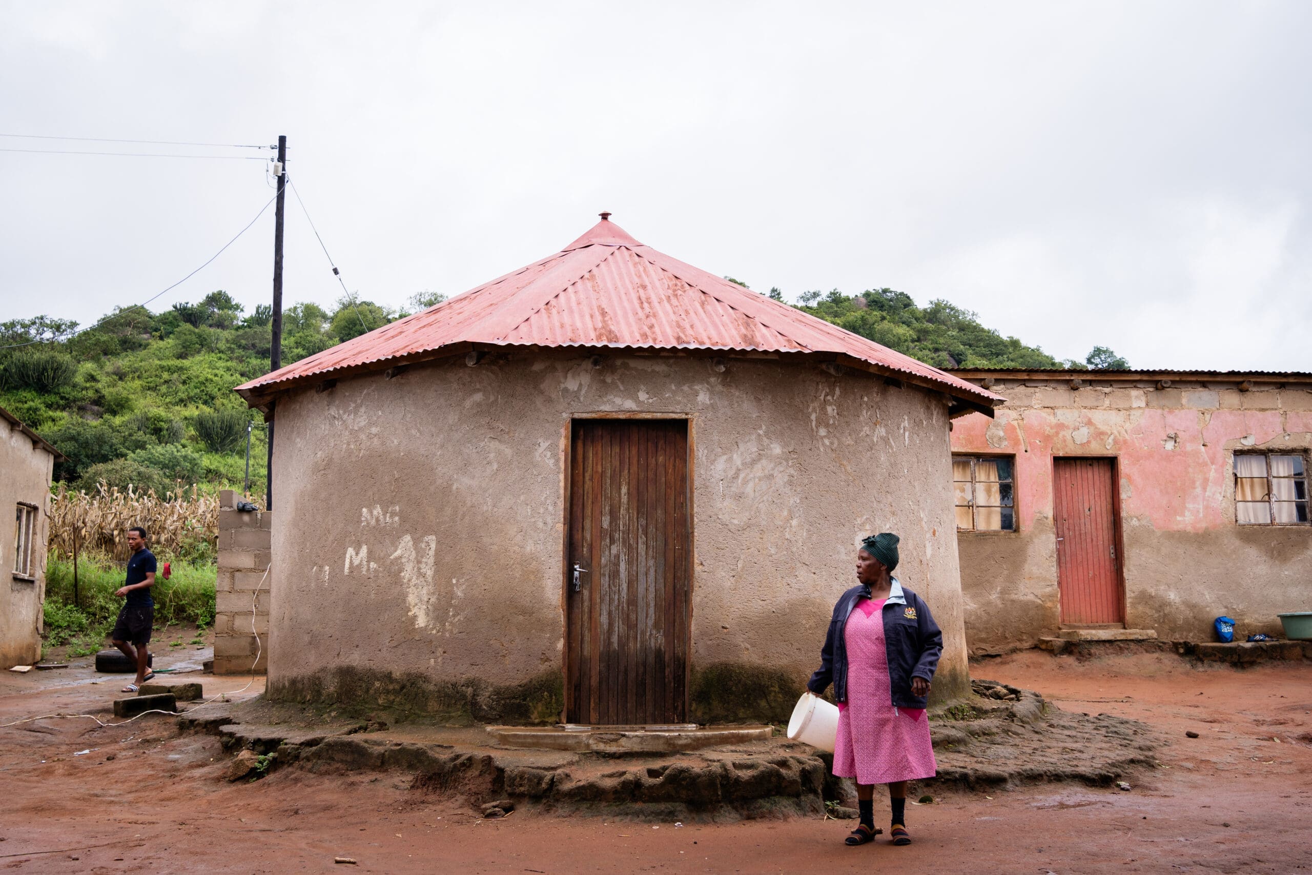 A woman in a pink dress stands outside a round, mud home with a red roof, holding a white container. Another person walks near a rectangular building with faded pink walls in the background. The area is muddy and rural.