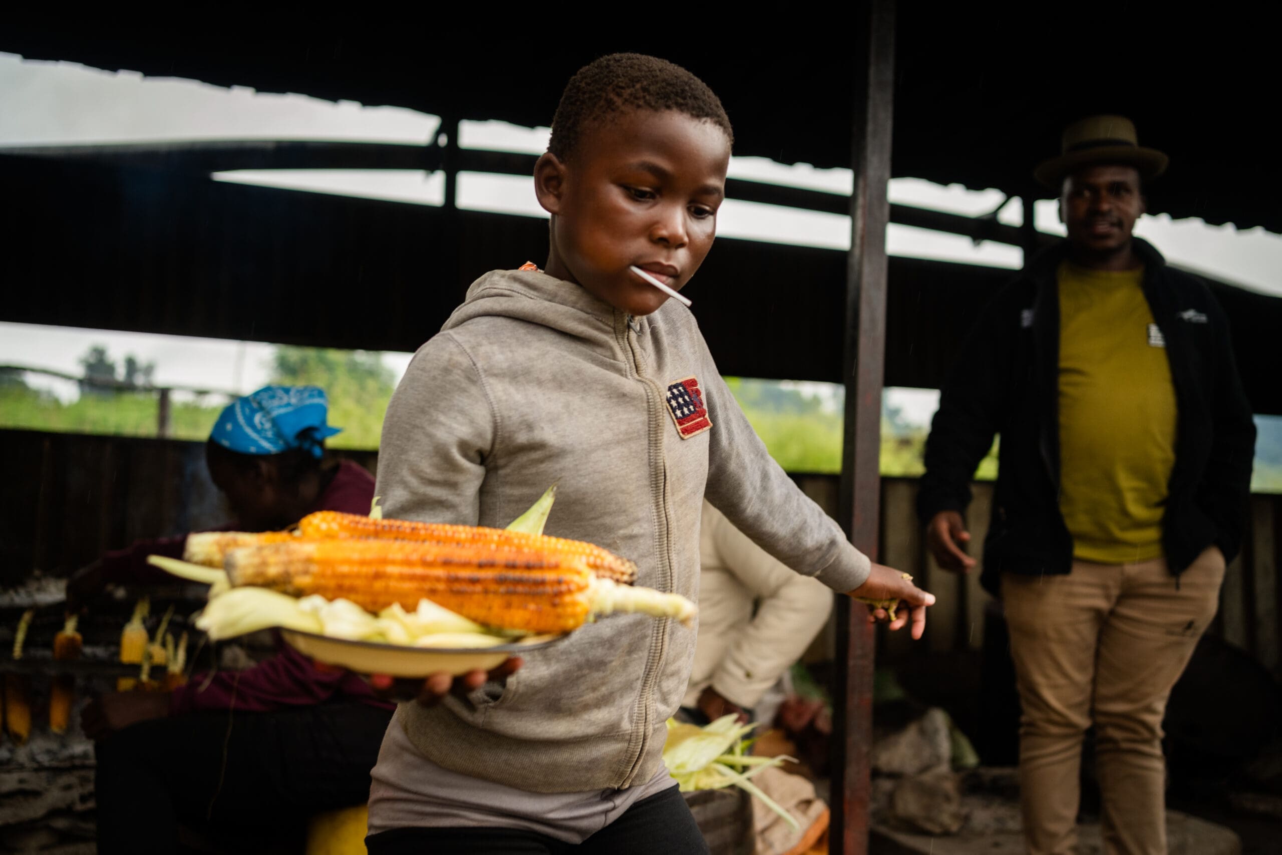 A young boy in a gray hoodie carries a plate with grilled corn. He has a stick in his mouth and looks focused. Two adults are in the background, one wearing a hat and another seated with a blue headscarf.
