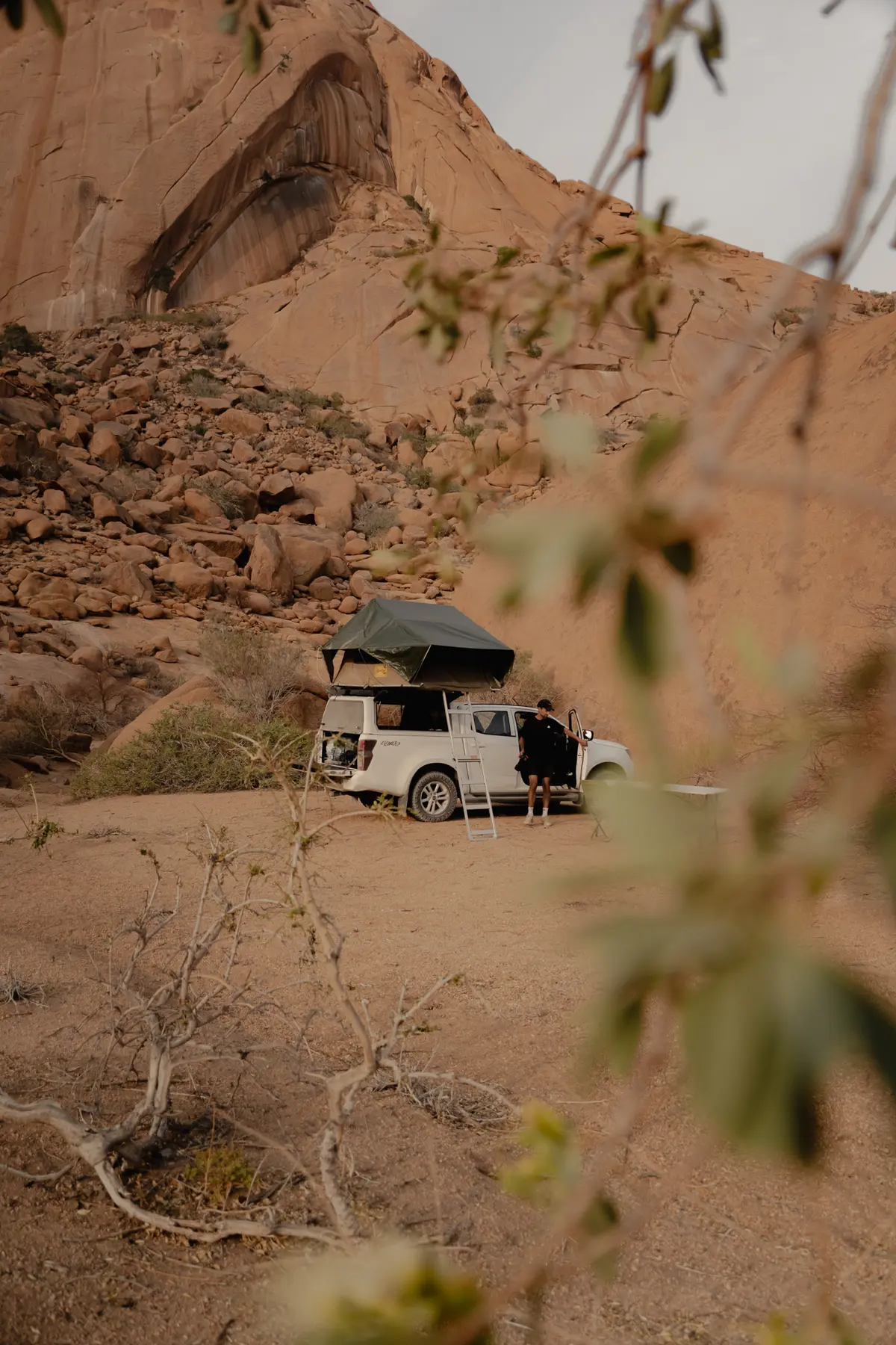 Een camper met een daktent staat geparkeerd in een rotsachtig woestijnlandschap. Een persoon staat naast het busje en op de achtergrond verrijzen grote zandsteenformaties. Op de voorgrond zijn vage takken zichtbaar.