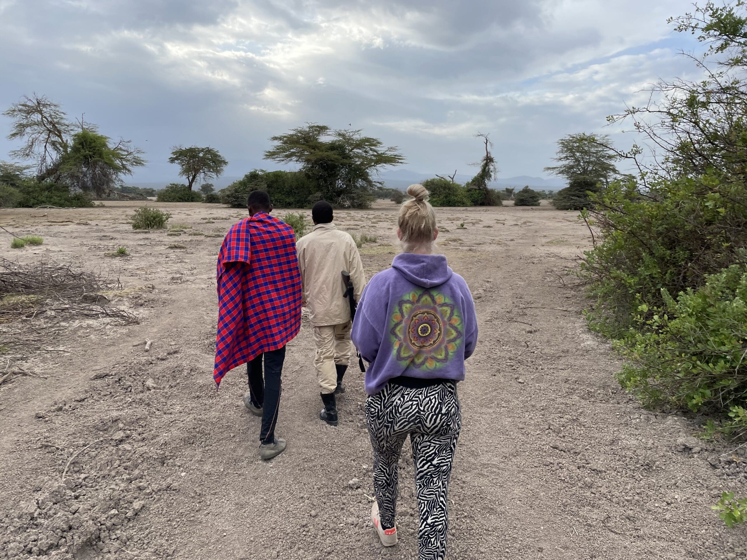 Three people walk on a dry, rocky landscape under a cloudy sky. One wears a red-checkered shawl, another wears beige, and the third wears a purple hoodie with a colorful design and zebra-print pants. Sparse trees are in the background.