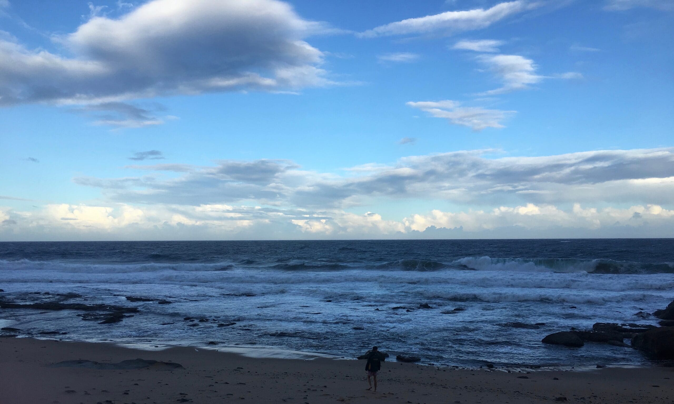Een persoon wandelt alleen op een zandstrand bij de kustlijn, met golven die beuken en een gedeeltelijk bewolkte blauwe lucht boven hem. De scène voelt kalm en weids aan.