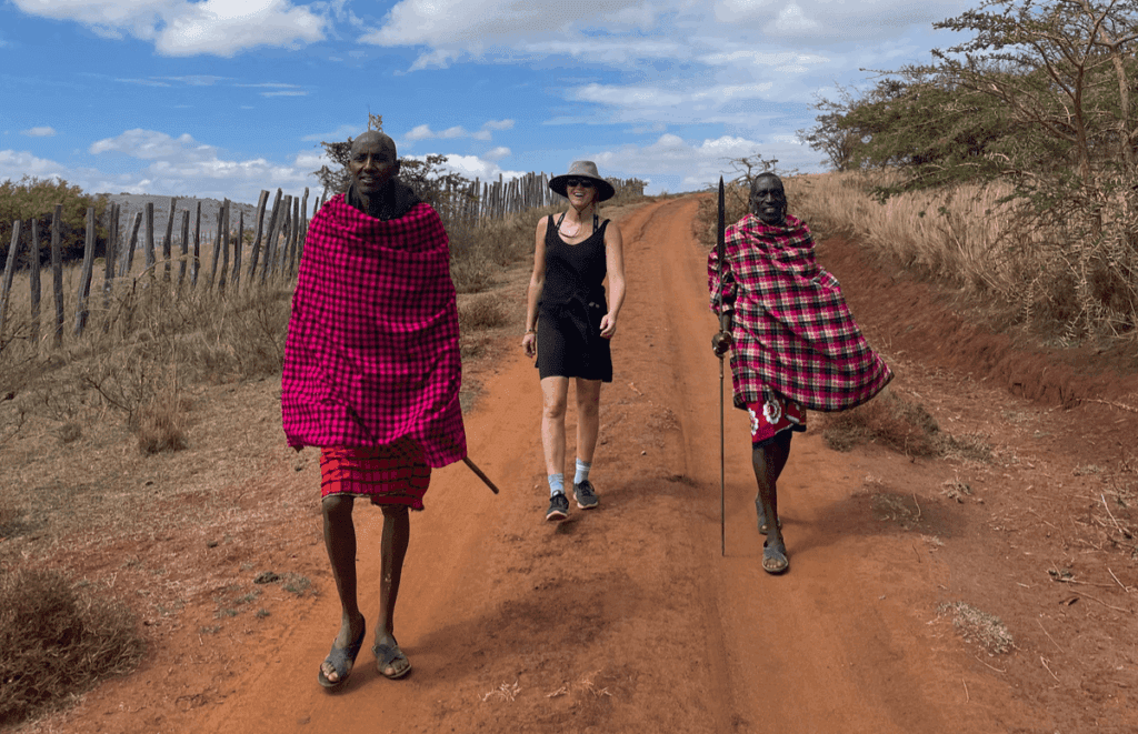 Three people walk on a red dirt road in Kenya; two Maasai men in red patterned shukas flank a woman in a black dress and hat. Travelling through Africa, they pass dry grass, wooden fences, and a partly cloudy sky.