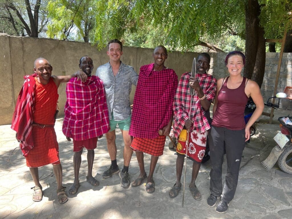 Fünf Maasai-Männer in traditioneller Kleidung und zwei lächelnde Touristen stehen an einem sonnigen Tag im Freien und posieren für ein Gruppenfoto im Schatten eines Baumes.