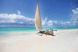 Een traditionele houten zeilboot met een beige zeil en meerdere mensen aan boord rust op de oever van een wit zandstrand in Tanzania, met turquoise water en een andere zeilboot in de verte onder een gedeeltelijk bewolkte hemel.