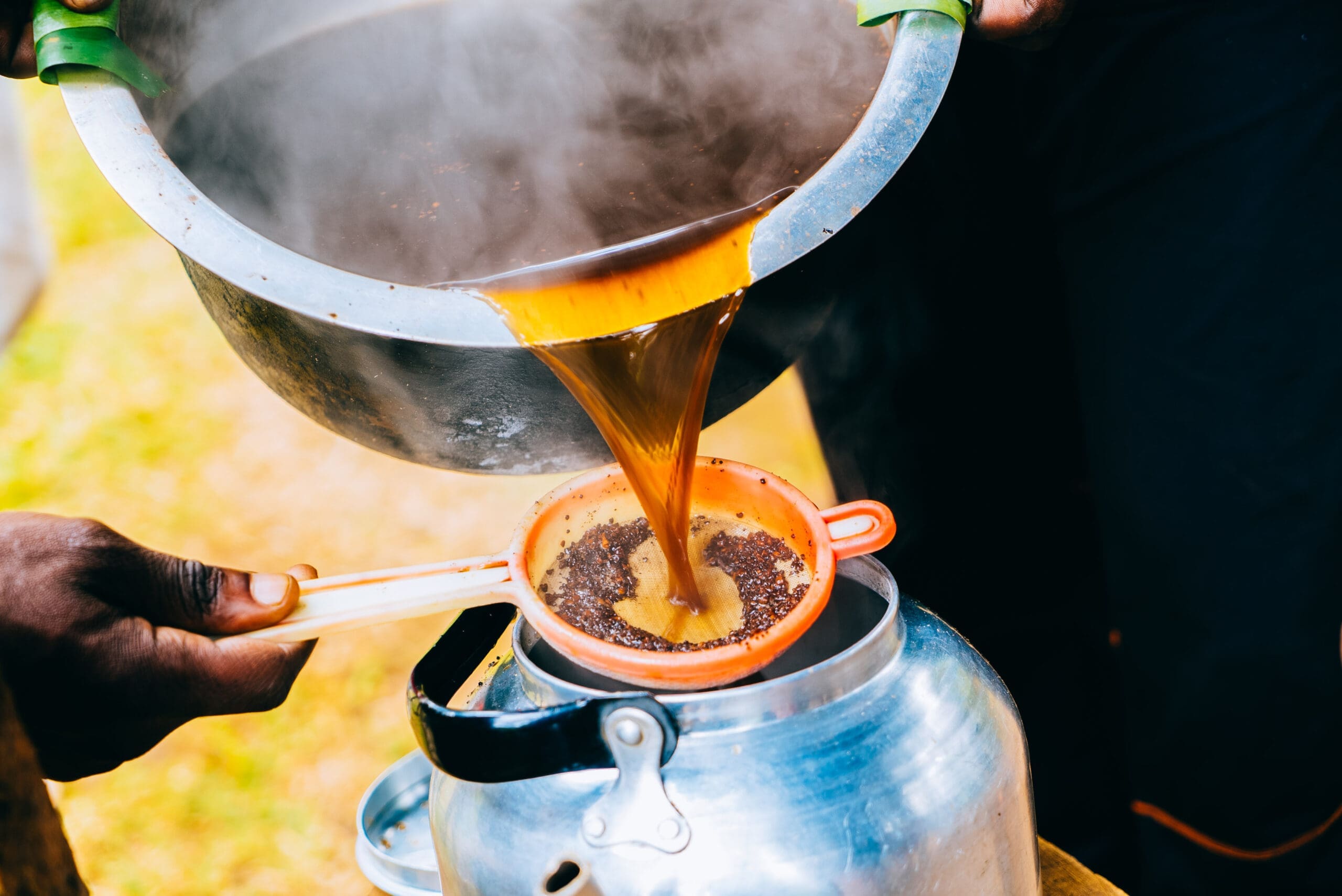 Een close-up van thee die vanuit een pot door een zeef in een ketel wordt gegoten, met stoom die opstijgt en handen die het keukengerei vasthouden, buiten in natuurlijk licht - de essentie vattend van traditionele theerituelen tijdens een reis door Tanzania.