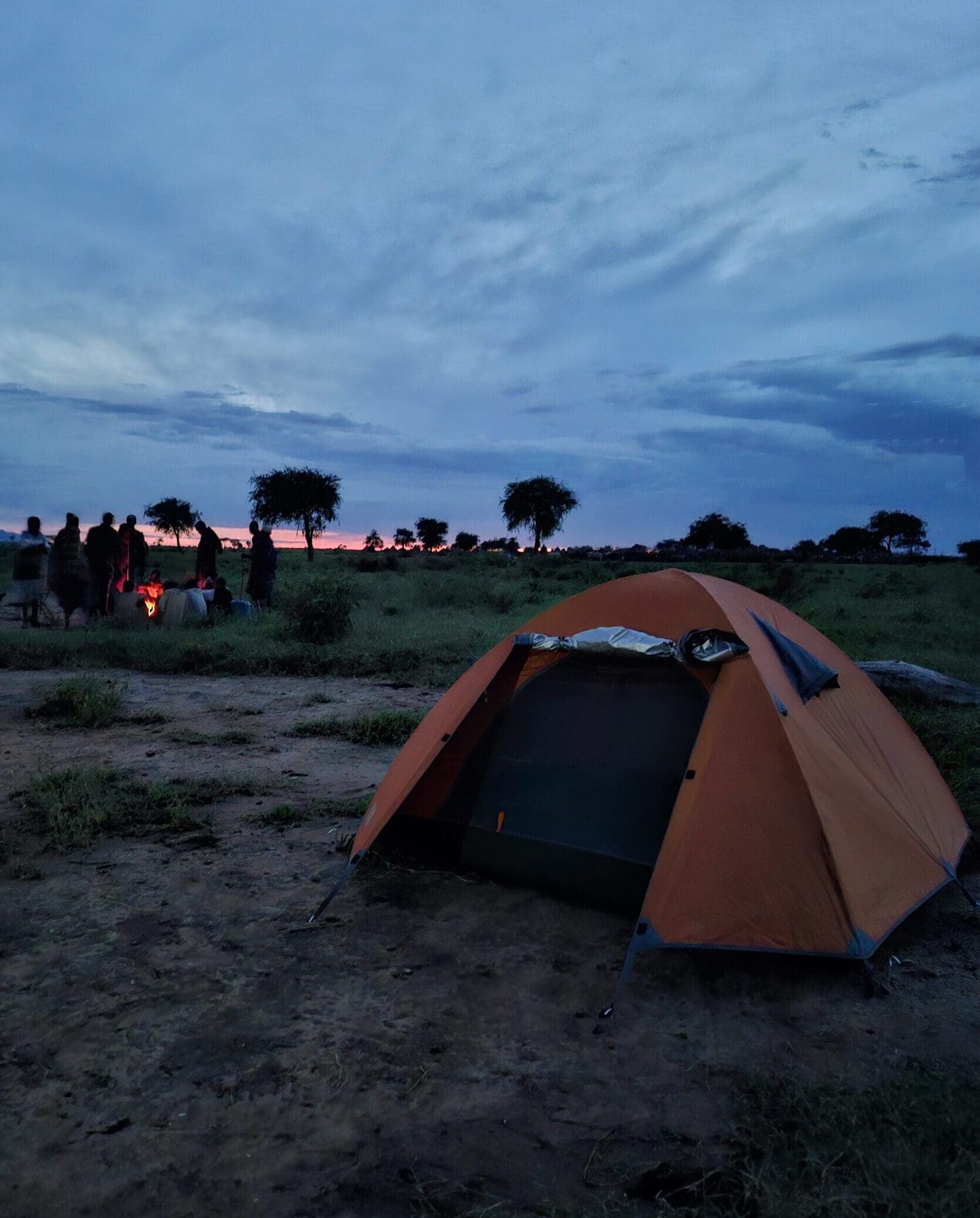 Een oranje tent is opgezet op een grasveld in de schemering. Op de achtergrond verzamelen reizigers zich rond een kampvuur onder een bewolkte hemel, met verspreide bomen in silhouetten tegen de horizon van Afrika.