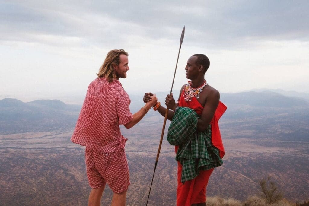 Ein Mann in moderner, rot gemusterter Kleidung grüßt einen traditionell gekleideten Massai-Stammesangehörigen, der einen Speer hält, vor einer malerischen Landschaft und Bergen im Hintergrund.