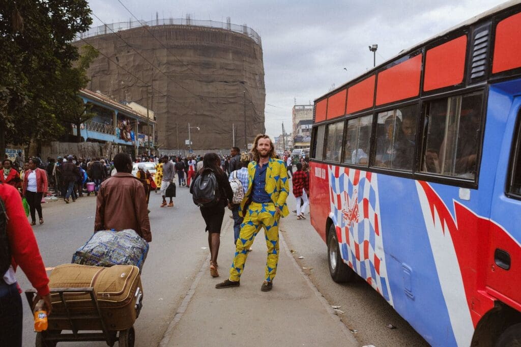 A man in a bright yellow and blue patterned suit stands on a busy city sidewalk near a colorful bus, surrounded by people travelling and carrying bags, with a large scaffolded building in the background, evoking the vibrant spirit of Kenya.