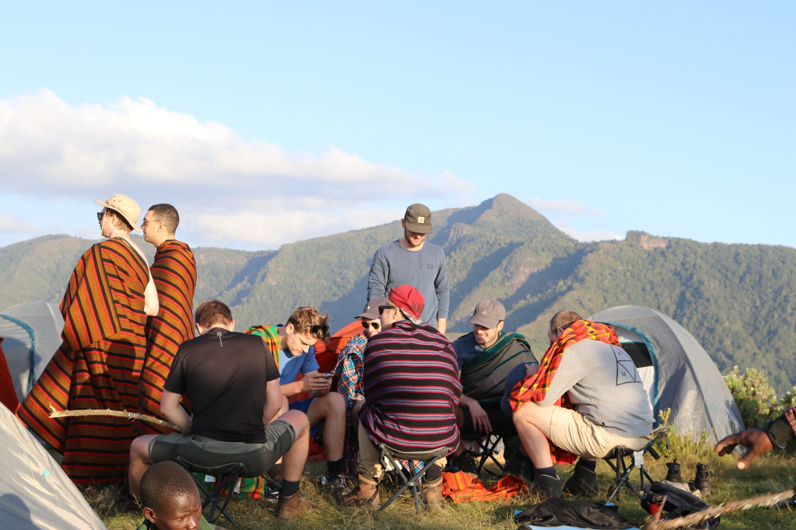 Een groep reizigers zit en praat tussen de tenten op een kampeerterrein in de bergen. De meesten dragen kleurrijke gestreepte sjaals, met op de achtergrond groene heuvels en een blauwe Oegandese lucht bezaaid met verspreide wolken.