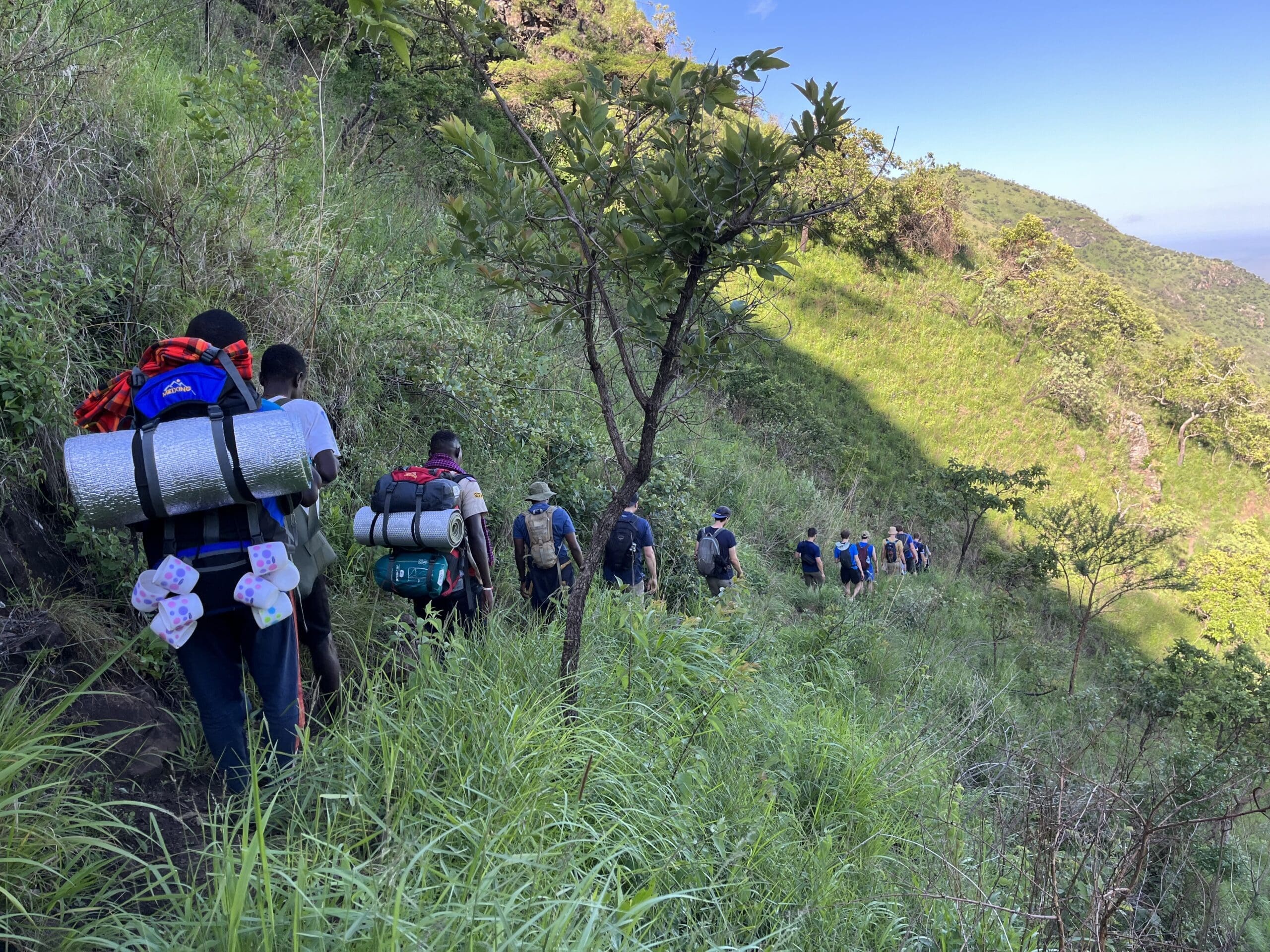 Een groep reizigers met rugzakken en kampeerspullen loopt achter elkaar langs een smal pad op een weelderige, groene heuvel in Oeganda onder een strakblauwe hemel.
