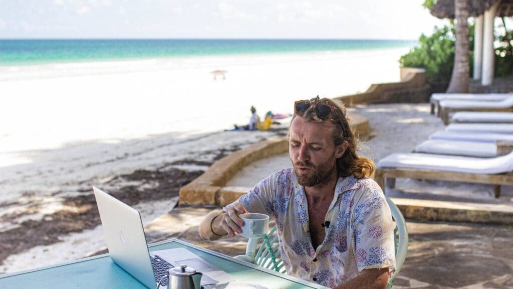 A man with long hair sits at a beachside table in Kenya, working on a laptop and holding a cup of coffee. The beach, sea, and lounge chairs capture the relaxed vibe of travelling along Africa’s beautiful coast.