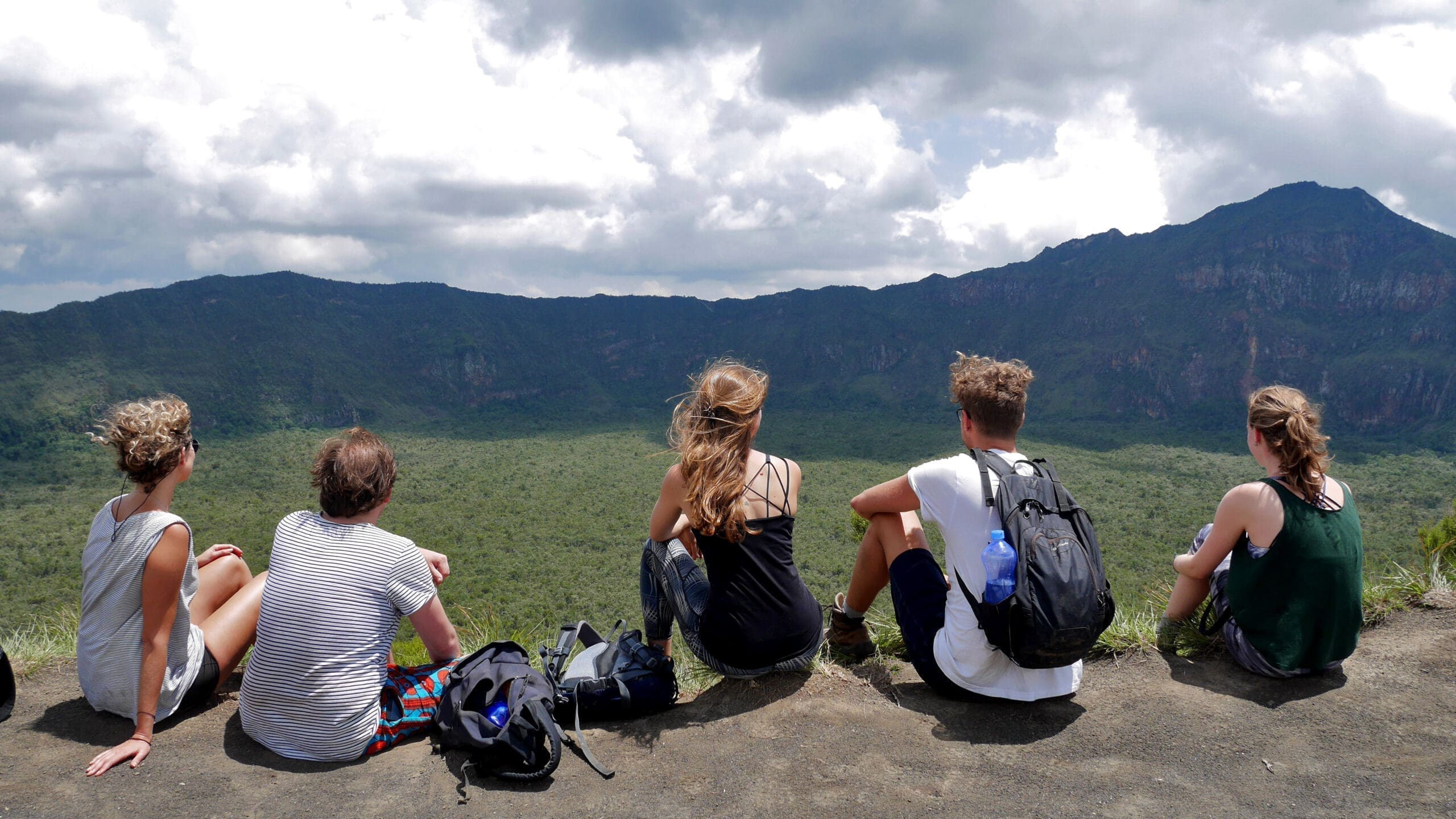 Five people sit on the edge of a cliff overlooking a vast green valley and distant mountains under a cloudy sky, with backpacks beside them.