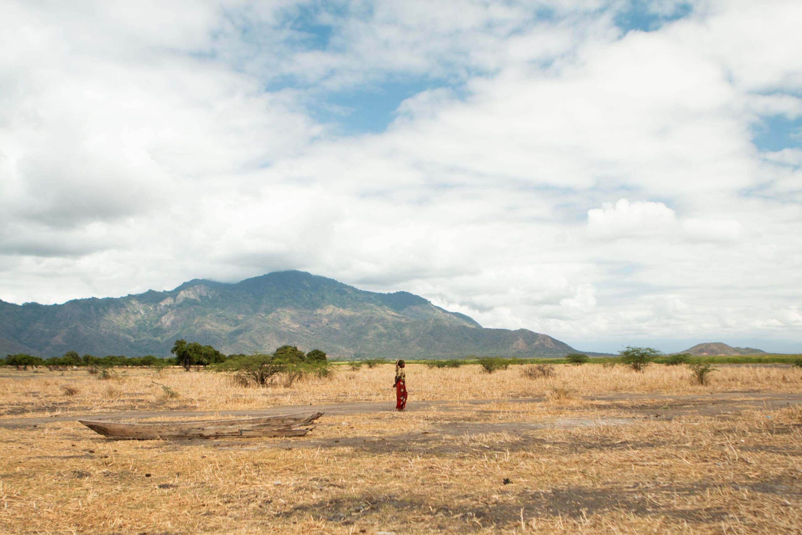 A single person in a red outfit stands in a vast, dry grassland with scattered trees, a wooden boat in the foreground, and mountains under a cloudy blue sky in the background.
