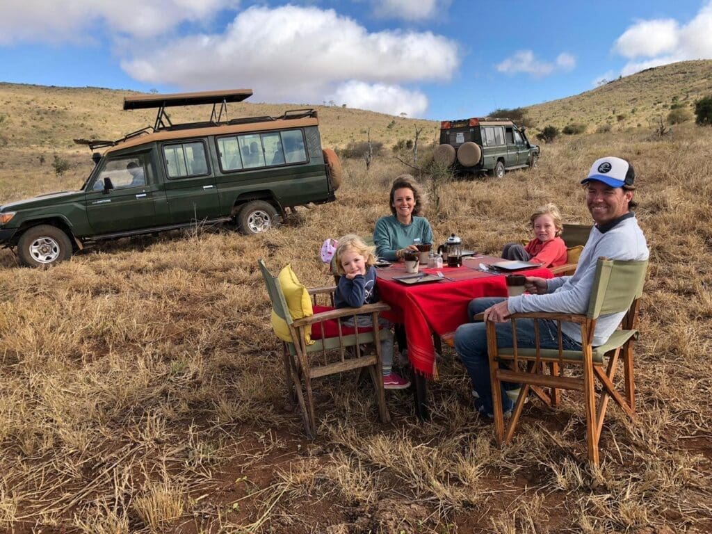Eine vierköpfige Familie sitzt an einem rot gedeckten Tisch und genießt eine Mahlzeit im Freien auf einer trockenen, grasbewachsenen Fläche. Zwei Safarifahrzeuge sind in der Nähe geparkt, und unter einem teilweise bewölkten Himmel sind sanfte Hügel zu sehen.