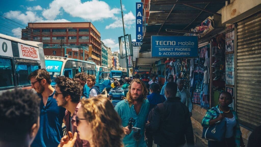 A busy urban street in Kenya with people walking, buses on the road, and shops selling phones and accessories. Colorful signs and crowded sidewalks create a lively city scene under a bright, partly cloudy African sky.