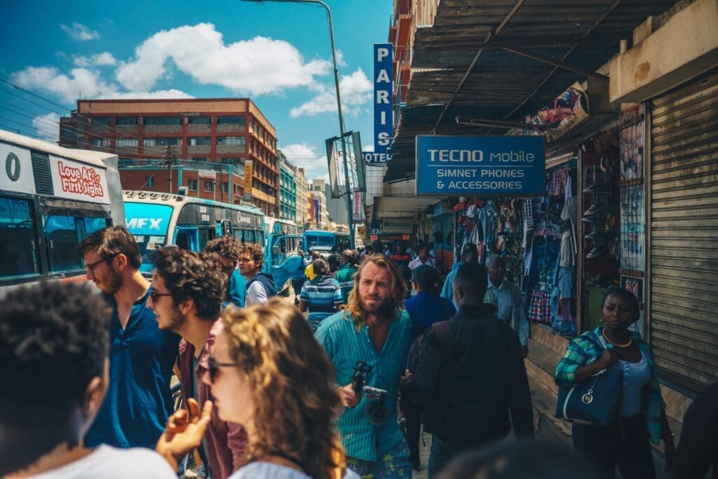 A busy street scene with many people walking, some looking at shops selling phones and accessories. Buses and cars fill the road, and buildings line both sides under a partly cloudy sky.