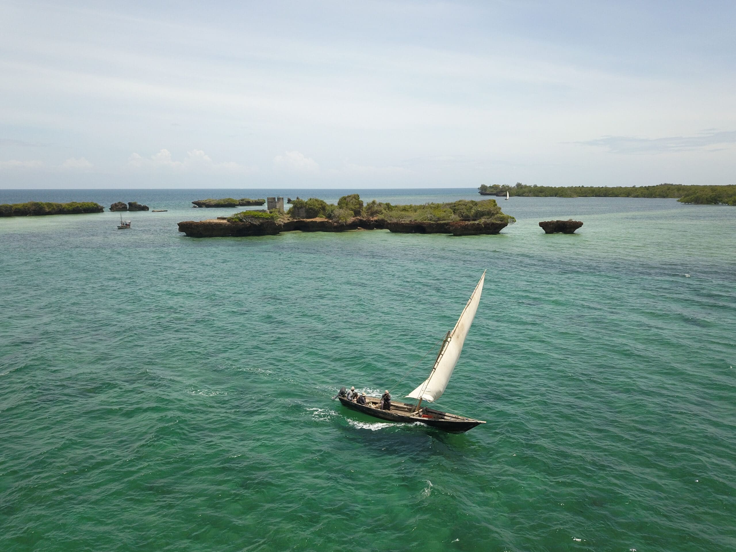 Een kleine zeilboot met mensen aan boord glijdt over turquoise water nabij rotsachtige, groene eilanden onder een heldere hemel.