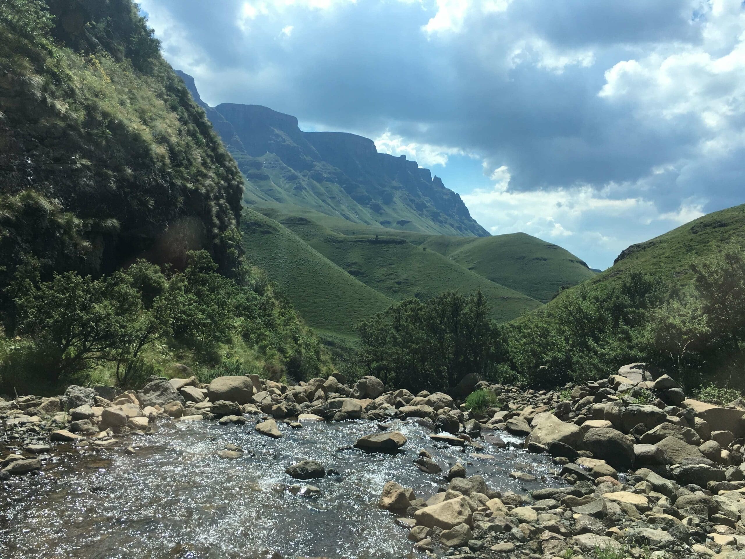 Een rotsachtige beek stroomt door een groene vallei, omzoomd door struiken en met gras begroeide heuvels, die reizigers uitnodigen om op verkenning te gaan. Torenhoge bergen rijzen op in de achtergrond onder een wolkenlucht, die de geest van het reizen in Zuid Afrika vastlegt.