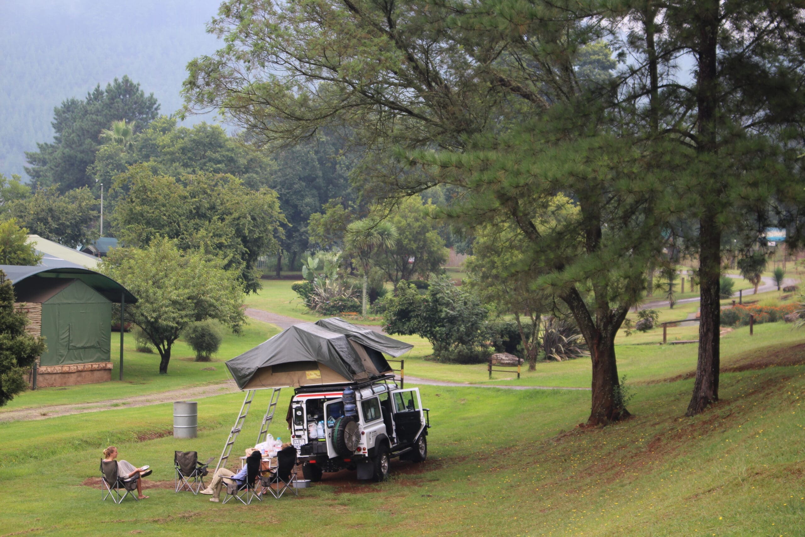 A group of people relax in camping chairs beside a white SUV with a rooftop tent in a grassy, wooded campsite surrounded by trees and greenery. Tents and forested hills are visible in the background.