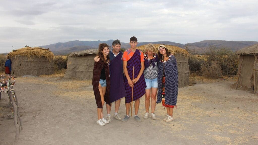 Four people stand smiling together in front of traditional huts, wearing colorful Maasai-style blankets. The background features dry grass, shrubs, mountains, and a cloudy sky.